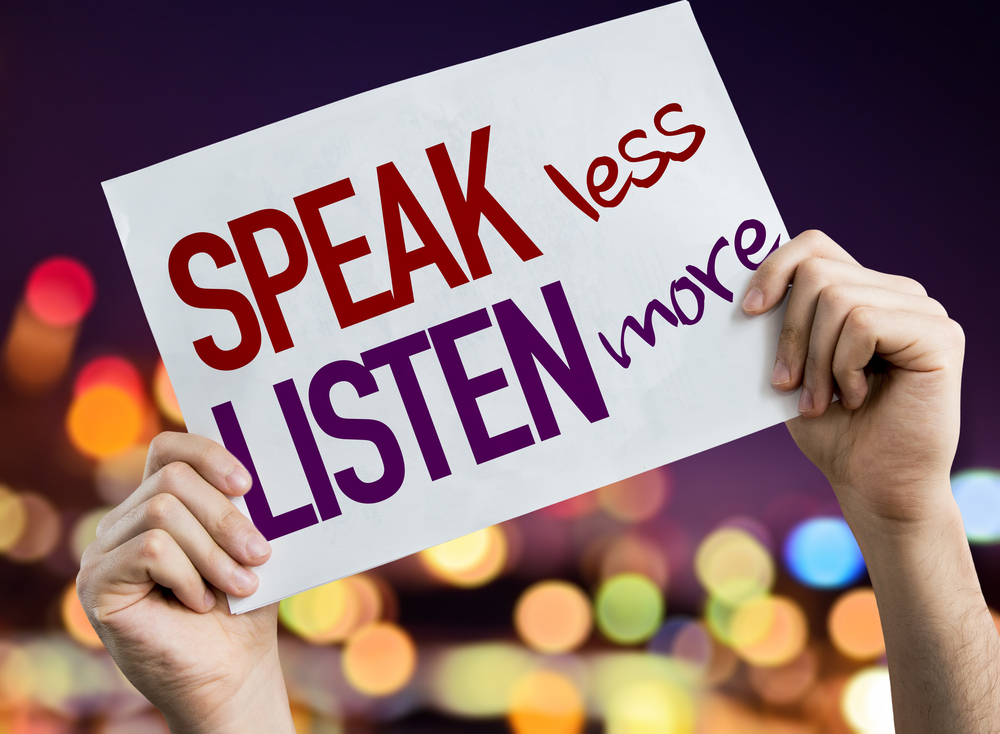 Speak Less Listen More placard with night lights in the background.